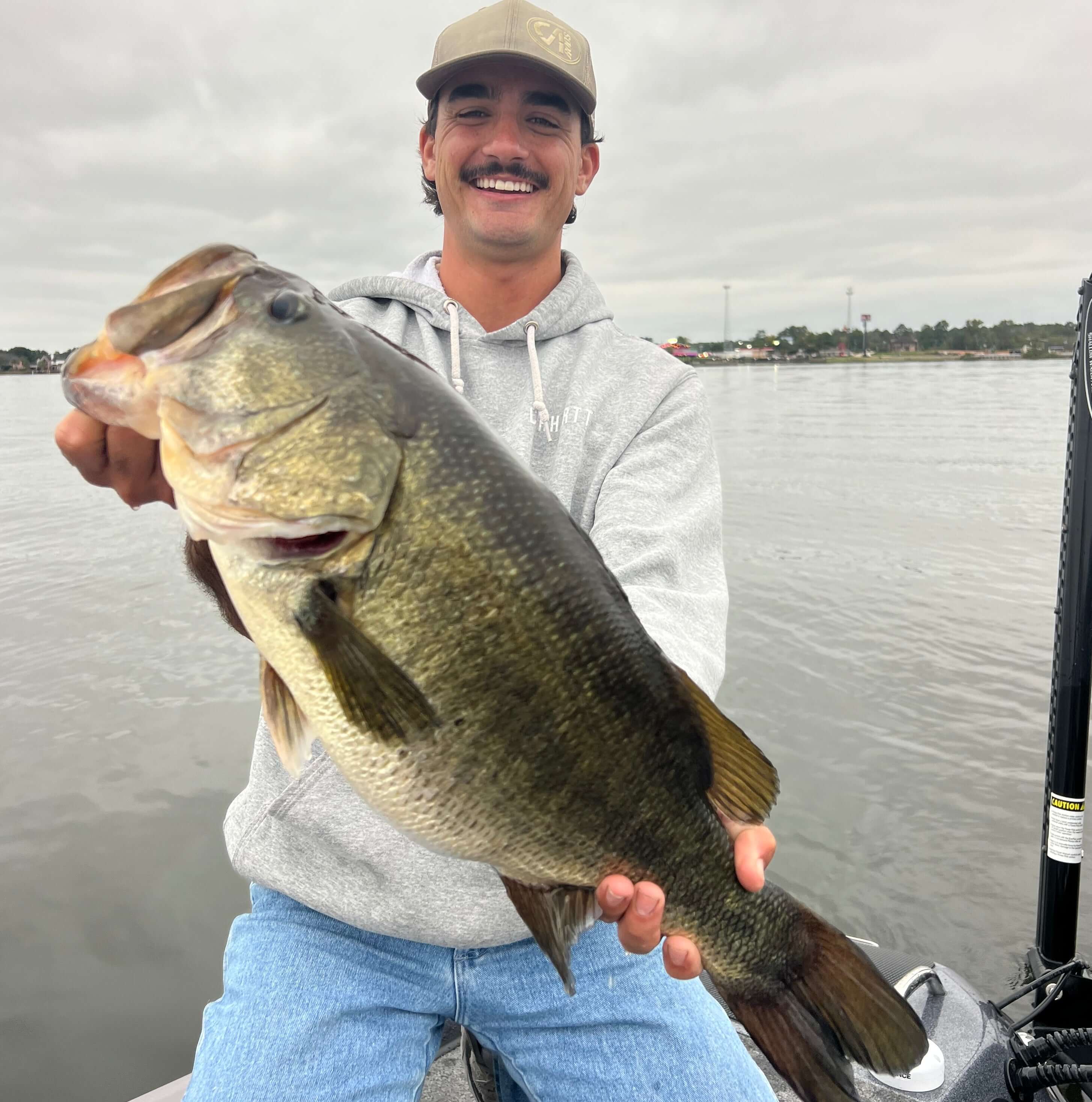 Fisherman proudly displaying a large bass catch on a cloudy day by the water.