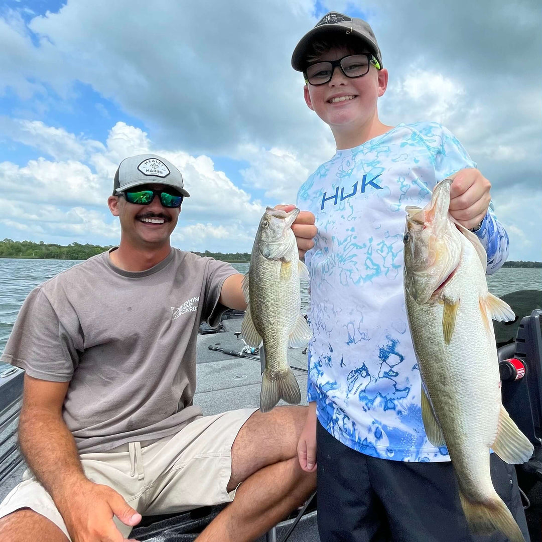 Father and son proudly holding up caught bass while fishing on a boat under a cloudy sky.
