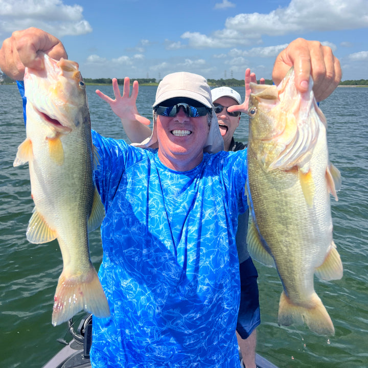 A man in a blue shirt holding up two largemouth bass with his friend behind him making a face. 