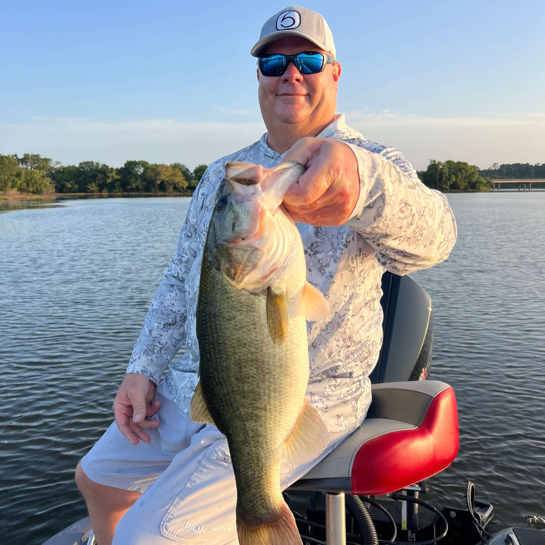 Man holding a large bass fish on a boat, smiling by the lake on a sunny day.