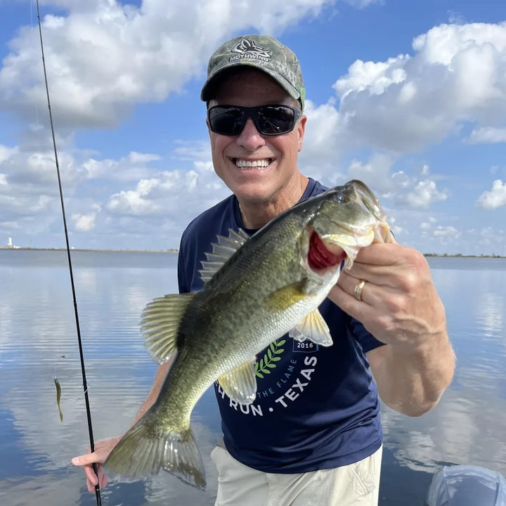 Angler smiling and holding a large bass fish while fishing on a sunny day by the water.