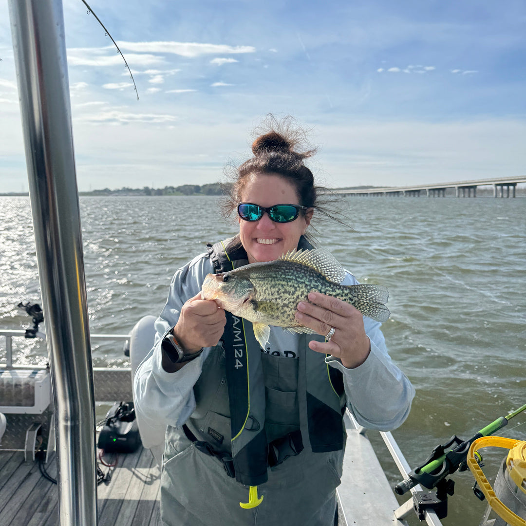 Woman wearing sunglasses holds a fresh caught crappie fish while fishing on a boat in a sunny lake.