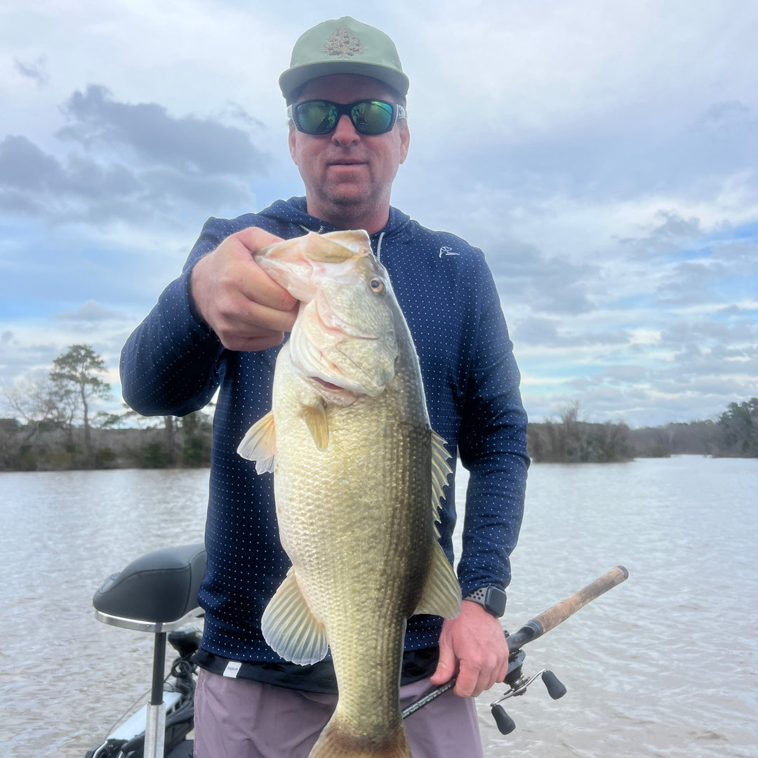 Angler holding a large bass fish, wearing sunglasses and a cap, with a scenic lake background.