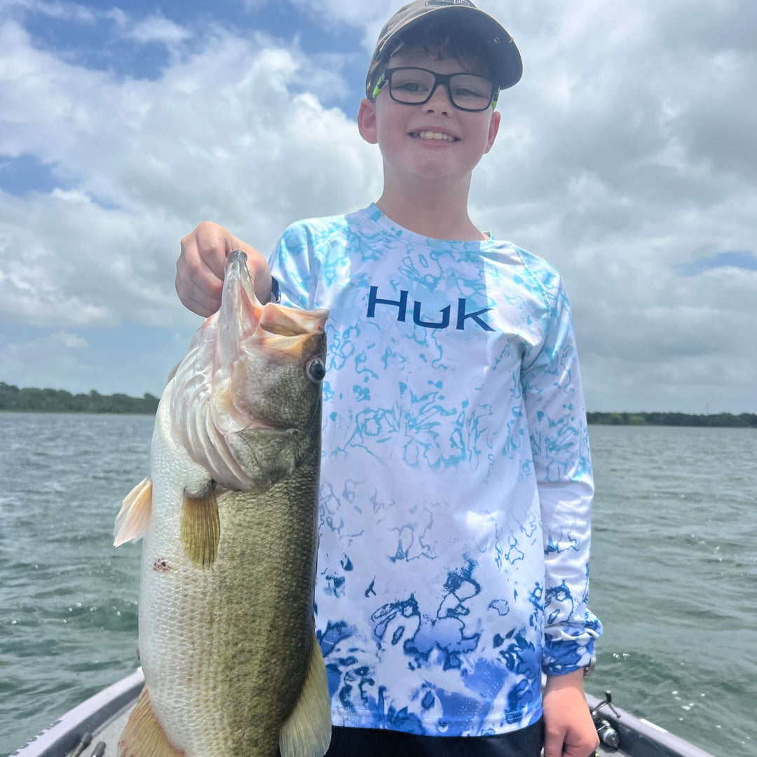 Young boy in a blue Huk fishing shirt proudly holding a large bass on a boat under a cloudy sky.